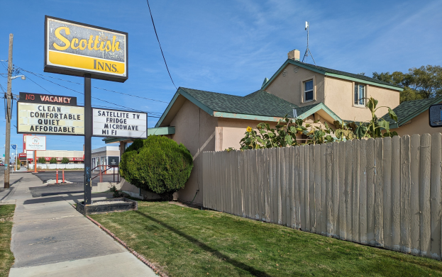 Ein schottisches Motel in San Francisco, Kalifornien, mit einem Haus mit Dach und Fenstern, einem Holzzaun, Pflanzen, Gras, Bäumen, Schildern, Strommasten mit Drähten, einer Straßenlaterne, Fahrzeugen auf der Straße und einer bewölkten Himmel.