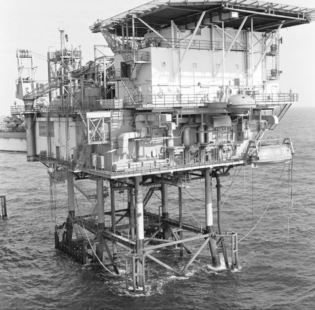 Ein Schwarz-Weiß-Foto einer Ölbohrinsel in der Mitte des Ozeans, mit einem Schiff im Hintergrund und dem Himmel darüber.