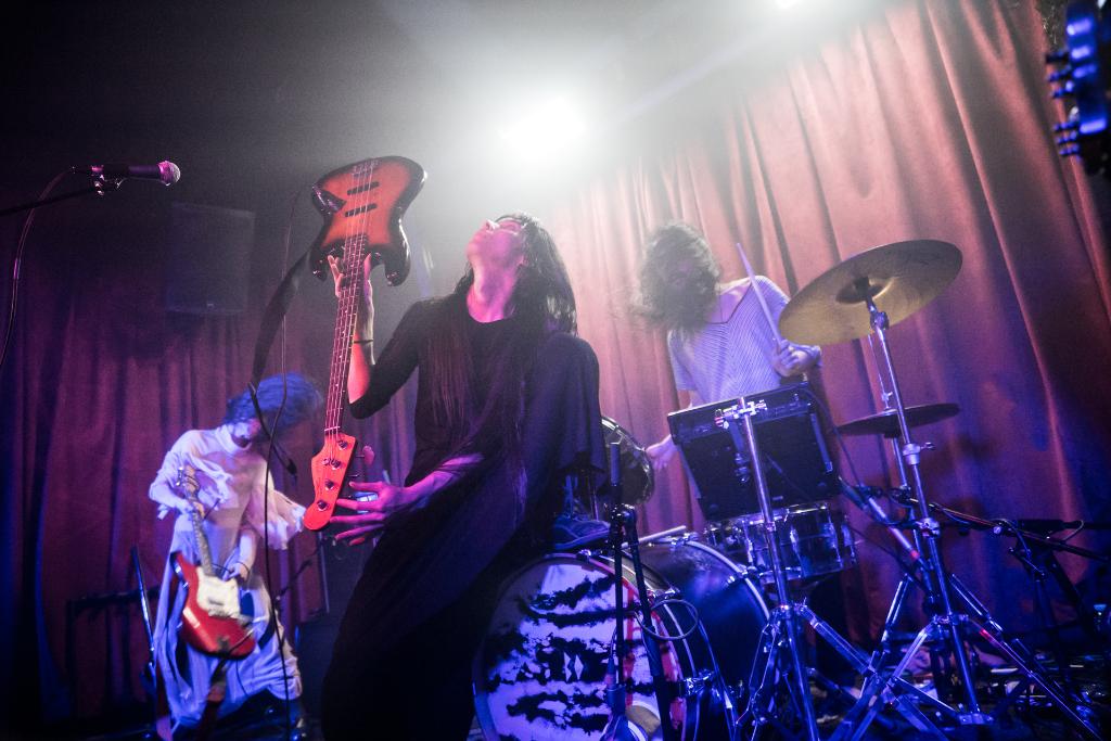 Eine Band performs auf der Bühne mit zwei Frauen, die links Gitarren spielen, einem Drummer im Hintergrund und einem Mikrofon vor den Gitarrenspielerinnen, sowie Bühnenlicht und Vorhängen.