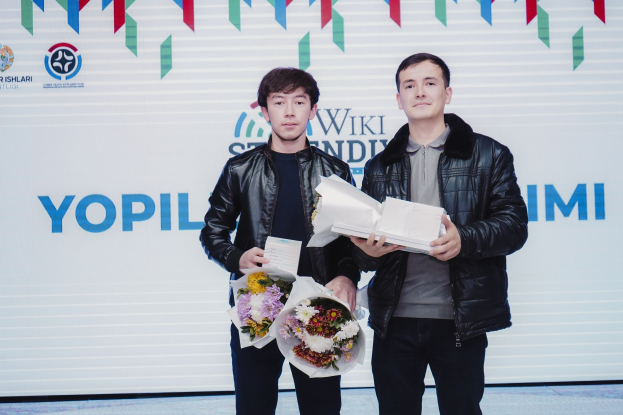 Zwei Männer in schwarzen Jacken mit Blumensträußen stehen auf einer Bühne mit einem weißen Banner im Hintergrund, auf dem 'Yopil International Film Festival' steht.