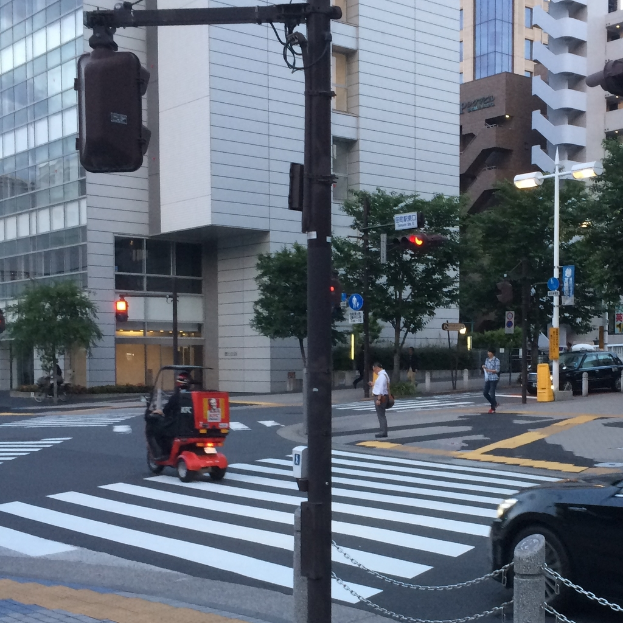 Ein Scooter ist auf der Straße mit einigen Personen im Hintergrund, eine Ampel im Vordergrund und Gebäude mit Bäumen in der Ferne.