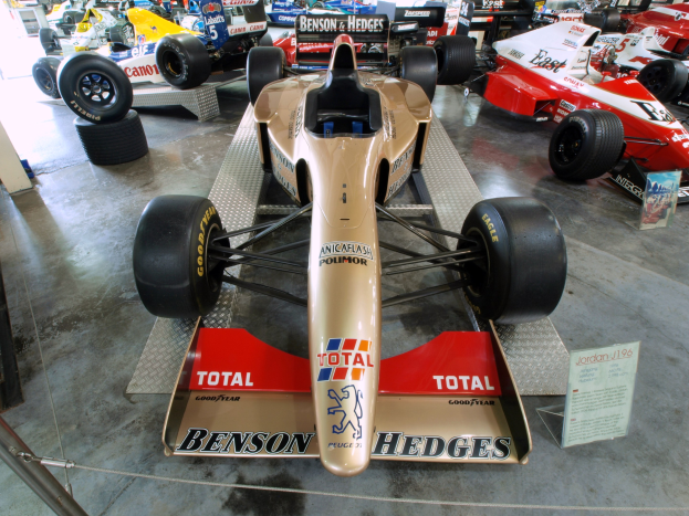 Rennwagen in einem Motorsportmuseum ausgestellt, getrennt durch einen Metallzaun von einer Tafel mit Text, mit Menschen und Beleuchtung im Hintergrund.