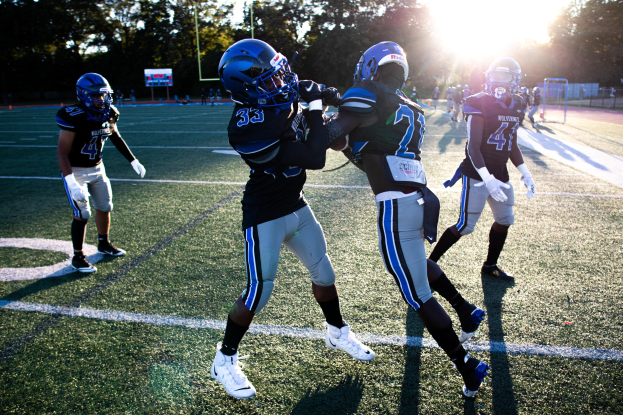 Gruppe junger Männer, die Fußball auf einem Feld mit Bäumen und einem klaren blauen Himmel spielen, tragen Helme und Schuhe.