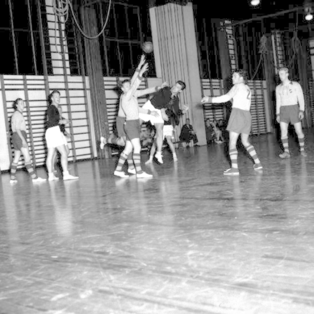 Eine Gruppe von Männern, die Handball auf einem Holzfußboden spielen, mit einer Wand und Lampen im Hintergrund, in Schwarz-Weiß.