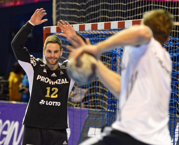 Zwei Männer spielen Handball vor einem Tor mit Netz und einem Banner mit Text im Hintergrund und einigen Menschen in einer Stadion.