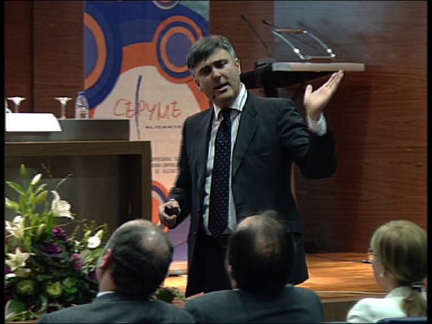 Zwei Männer und eine Frau sitzen auf Stühlen, während ein Mann in einem schwarzen Blazer an einem Podium mit Mikrofonen steht und in eines spricht, mit einem Blumentopf oder einem Blumenstrauß in der Nähe und einem Tisch mit einem Buch, einer Wasserflasche und anderen Gegenständen daneben, vor einer braunen Wand und einer bunten Tafel mit Text im Hintergrund, wahrscheinlich in einem Konferenzsaal.