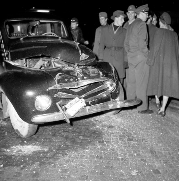 Ein schwarz-weißes Bild eines beschädigten Autos am Straßenrand, umgeben von einer Gruppe besorgter Menschen.