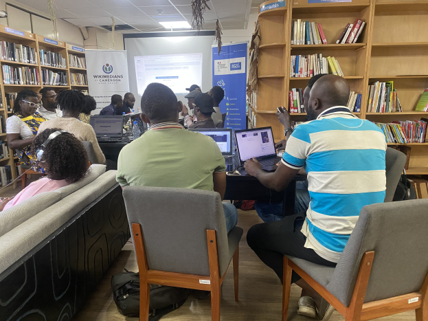 Gruppe von Menschen an einem Tisch mit Laptops umgeben von Bücherregalen, mit einem Banner, Bildschirm, Deckenleuchten und Taschen im Hintergrund.
