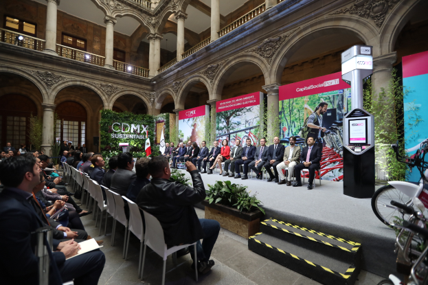 Eine Gruppe von Menschen sitzt auf Stühlen und schaut auf einen Mann auf einer Bühne, der ein Mikrofon hält, während Fahrräder rechts geparkt sind und verschiedene Elemente wie Plakate, Pflanzen, Säulen, Geländer, Lampen, Fenster und eine Wand im Hintergrund zu sehen sind.