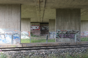 Eine Schienenstrecke mit Graffiti an der Seite, umgeben von Steinen, Gras, einem Zaun, Säulen, Drähten und einem Dach.