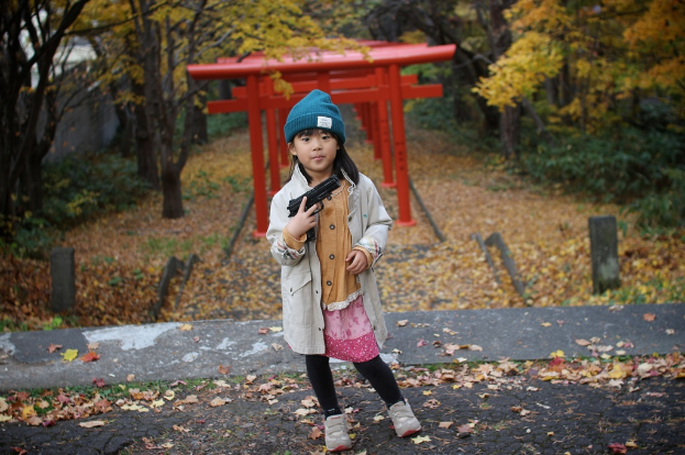 Ein Mädchen steht auf einer Straße und hält eine Waffe, trägt eine graue Jacke, mit Bäumen und roten Metallobjekten im Hintergrund und trockenen Blättern auf dem Boden.