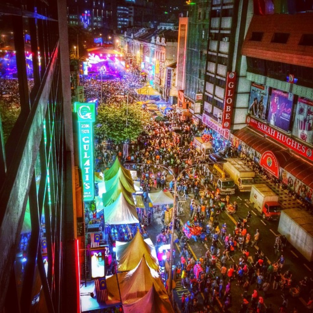 Eine volle KarnevalsSzene mit Zelten, Geschäften, Gebäuden und Bannern, mit hell erleuchteter Fläche im Hintergrund.