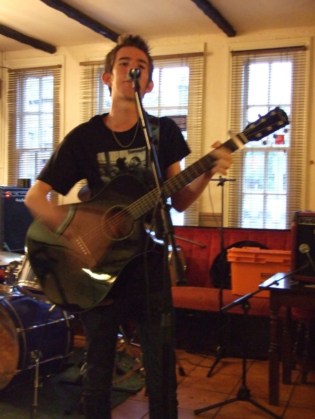 Ein Mann in schwarzem T-Shirt und Hose steht mit einer Gitarre und einem Mikrofon auf einem Ständer, mit einer blauen Trommel links im Bild, einer Couch mit Gegenständen darauf und durchsichtigen Vorhängen im Hintergrund unter einer Decke.