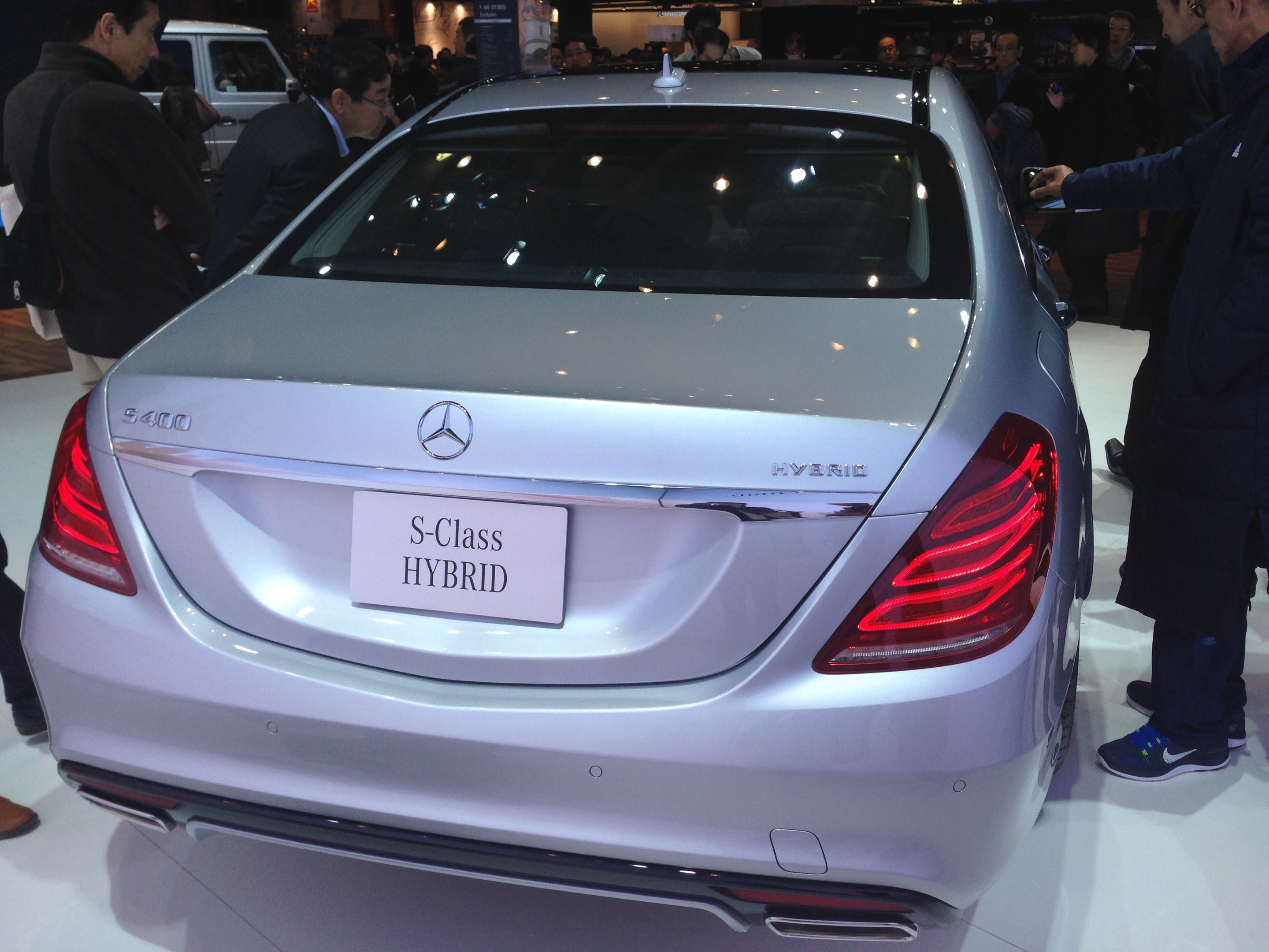 Silberner Mercedes S-Klasse Hybrid auf einer Automesse gezeigt, umgeben von Menschen in verschiedenen farbigen Kleidern, mit anderen Fahrzeugen und Beleuchtung im Hintergrund.