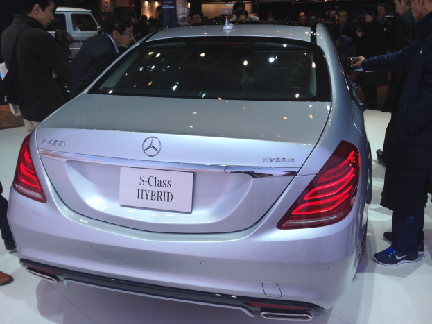 Silberner Mercedes S-Klasse Hybrid auf einer Automesse gezeigt, umgeben von Menschen in verschiedenen farbigen Kleidern, mit anderen Fahrzeugen und Beleuchtung im Hintergrund.