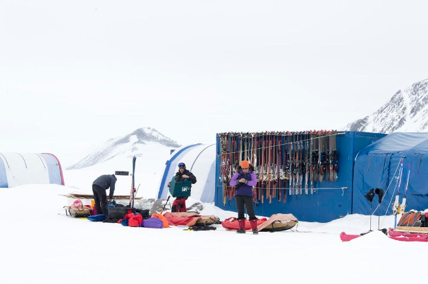 Drei Personen stehen auf einer verschneiten Landschaft mit verstreuten Taschen, Zelten mit Skiern dahinter und schneebedeckten Hügeln im Hintergrund unter einem klaren Himmel.