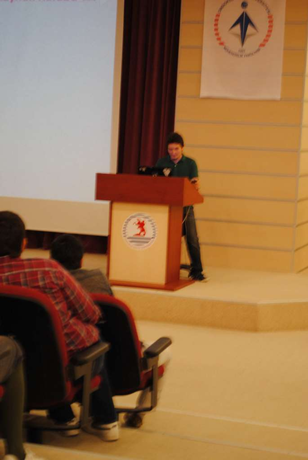 Eine Person steht auf einem Podium in der Mitte und spricht zu einem links sitzenden Publikum, mit einer Tafel und einem Vorhang links und einer weiteren Tafel im Hintergrund