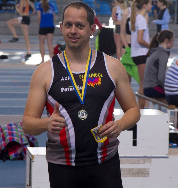 Ein Mann in einem schwarzen Trikot und einer Medaille steht, während im Hintergrund einige Menschen trainieren.