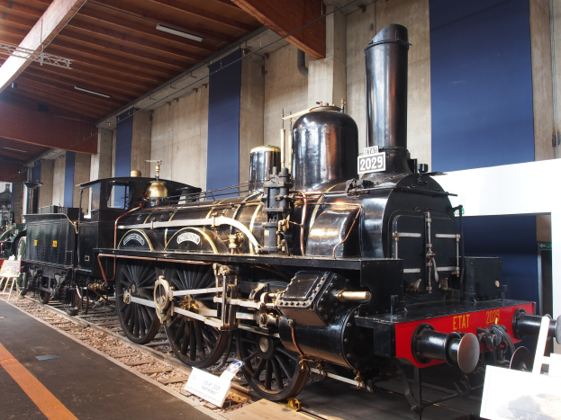 Eine große, alte Dampflokomotive auf Schienen in einem Museum ausgestellt, mit Informationsschildern, Ständen und anderen Ausstellungsstücken in der Nähe, vor einer Wand und Decke mit Beleuchtung.