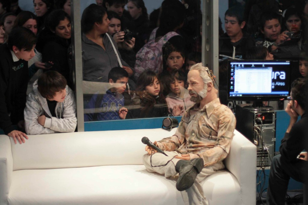 Eine Gruppe von Menschen, die ein Puppentheater fotografieren, wobei eine Person einen Bildschirm ansieht und eine puppenähnliche Person auf einem Sofa sitzt und ein Mikrofon hält.