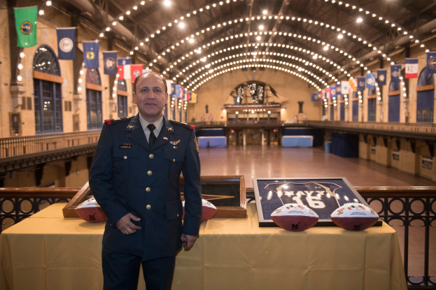Ein Mann in Uniform, identifiziert als Stabschef der National Football League (NFC), steht neben einem Tisch mit zwei Footballs und einem Foto Rahmen, mit Flaggen, Lichtern und anderen Gegenständen im Hintergrund.