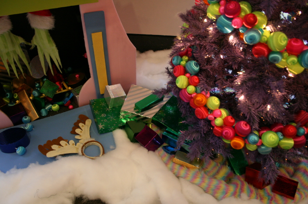 Drinnen steht rechts ein geschmückter Weihnachtsbaum mit Lichterkette und Schmuck, in der Mitte Geschenkboxen und Dekorationen, eine Hütte mit Dekorationen oben links und Baumwolle und Wolle unten.