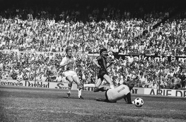 Schwarzes und weißes Foto von Fußballspielern im Einsatz auf einem Feld mit Zuschauern im Hintergrund und einer "Bundesliga 1978-79 Ajax 1-1 Hannover 96"-Fahne.
