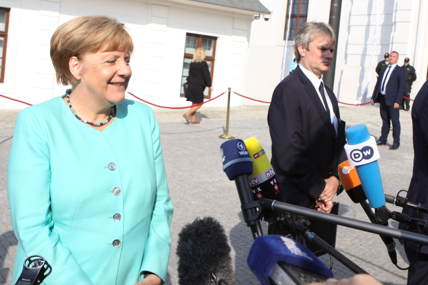 Bundeskanzlerin Angela Merkel spricht zu Reportern im Freien, umgeben von einer Gruppe mit Mikrofonen und einer Absperrung, unter einem bewölkten Himmel.