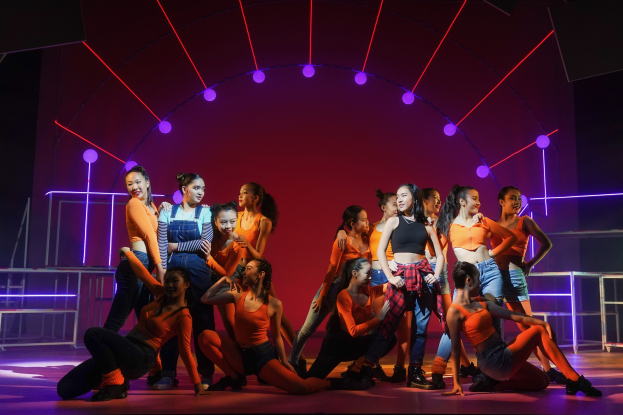 Eine Gruppe junger Tänzer in bunten Kostümen performt auf einer Bühne mit Geländern, beleuchtet von bunten Lichtern, mit einem jubelnden Publikum im Hintergrund.