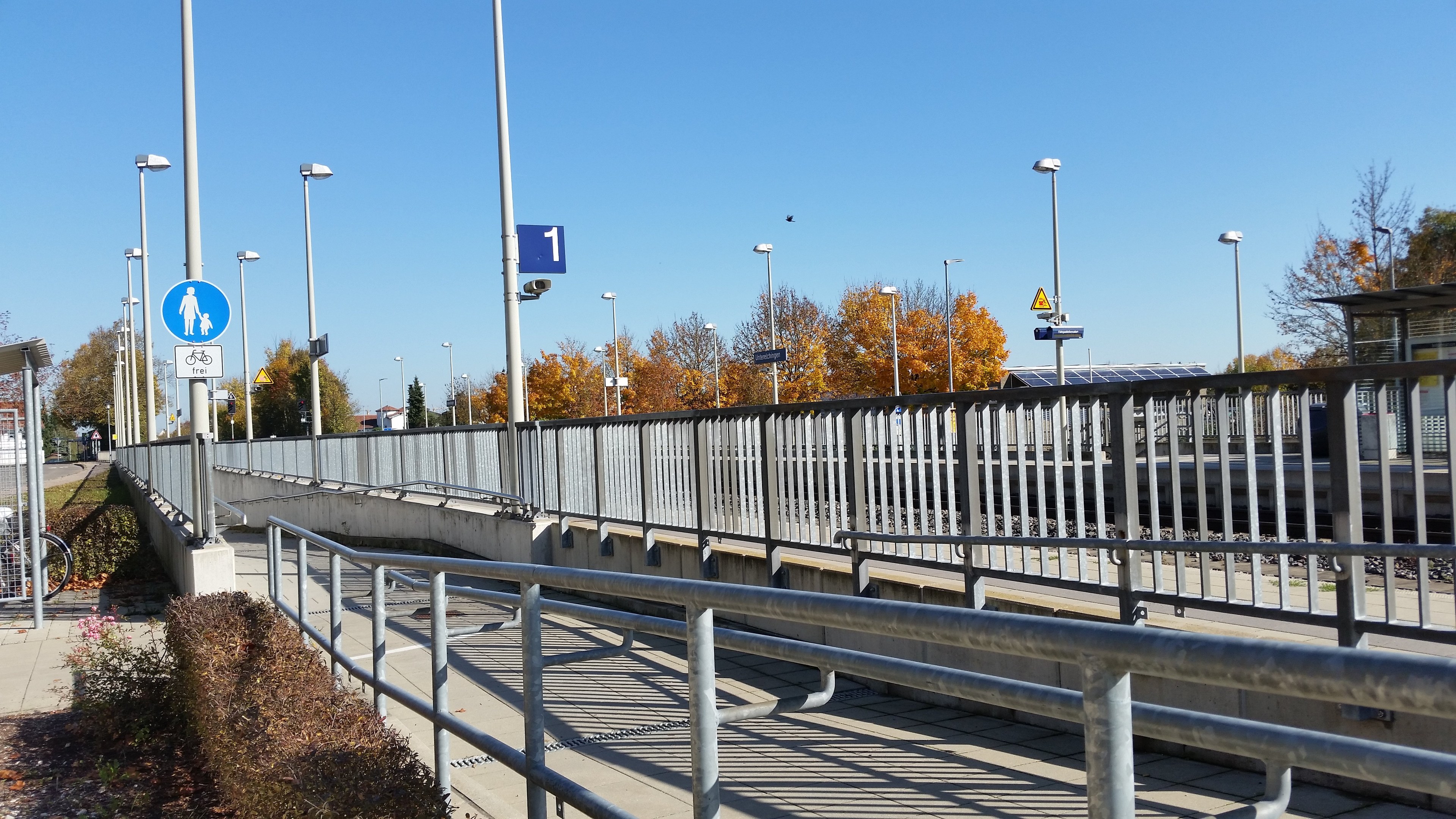 Eine Brücke mit Geländern, Laternen, Schildern, Fahrrädern, Pflanzen, Bäumen und einem Himmel als Hintergrund, mit einem parkenden Fahrrad an der Seite.