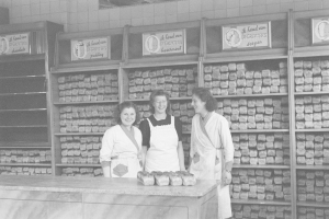 Drei Frauen in Schürzen lächeln vor einem Bäckertisch mit Lebensmitteln, Regalen und Wandtafeln im Hintergrund, dargestellt in Schwarz-Weiß.