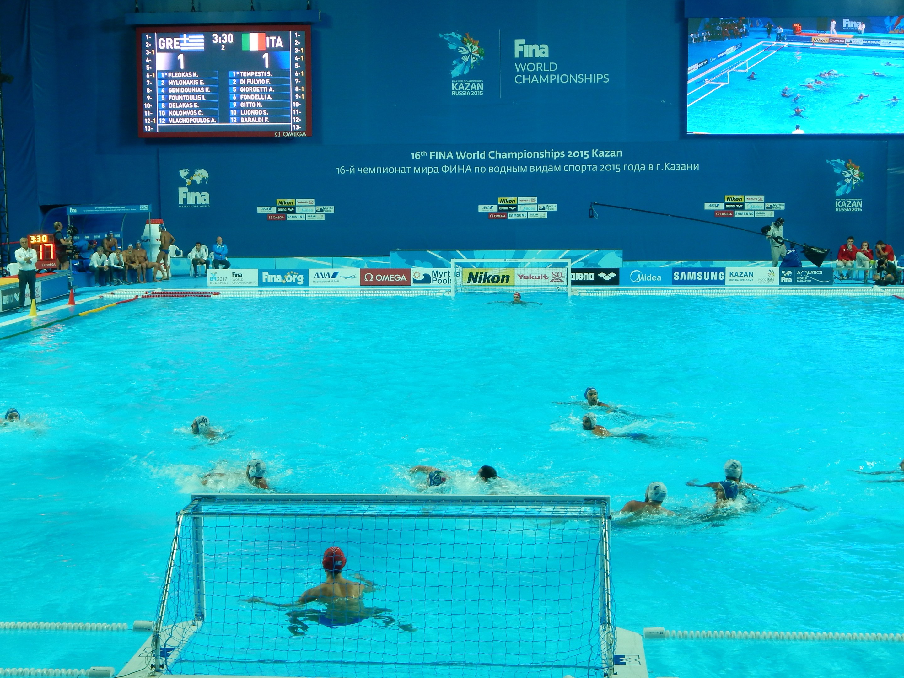 Gruppe von Athleten beim Wasserballspielen in einem Pool während der FINA-Weltmeisterschaften 2016 in Kazan, mit Zuschauern auf der linken Seite und einem Bildschirm im Hintergrund mit Event-Branding.