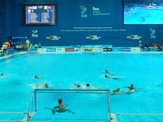 Gruppe von Athleten beim Wasserballspielen in einem Pool während der FINA-Weltmeisterschaften 2016 in Kazan, mit Zuschauern auf der linken Seite und einem Bildschirm im Hintergrund mit Event-Branding.
