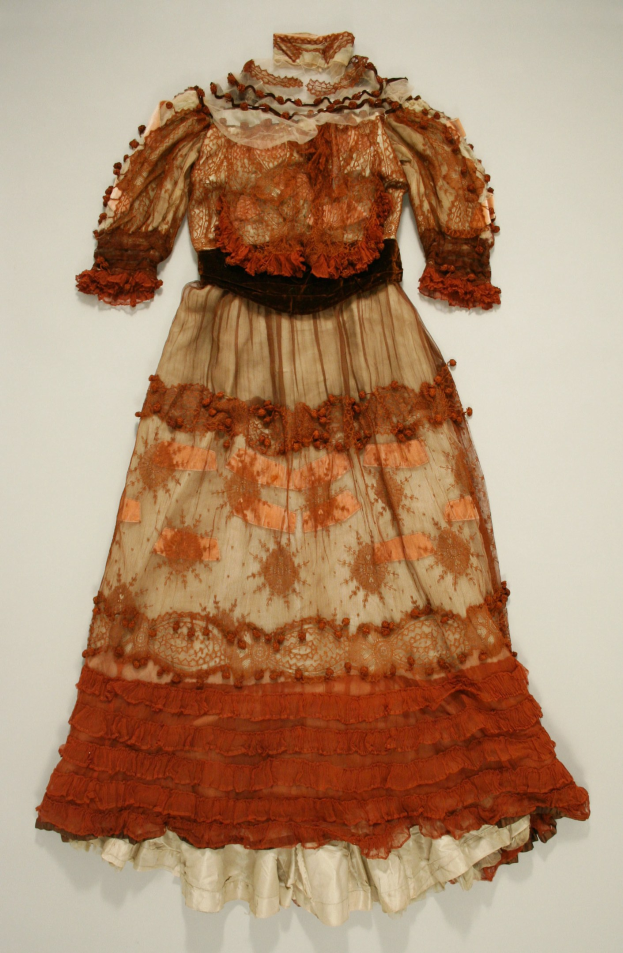 Ein helles Kleid mit hohem Ausschnitt, langen Ärmeln und einem bodenlangen Rock, das auf einem Schaufensterpuppen gegen einen weißen Hintergrund in einem Museum präsentiert wird.