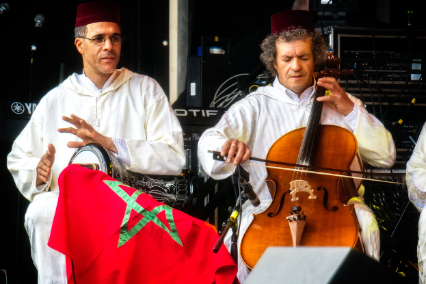 Zwei Männer in passenden Kostümen und Mützen sitzen und spielen verschiedene Musikinstrumente, hinter ihnen sind Musiksysteme zu sehen.
