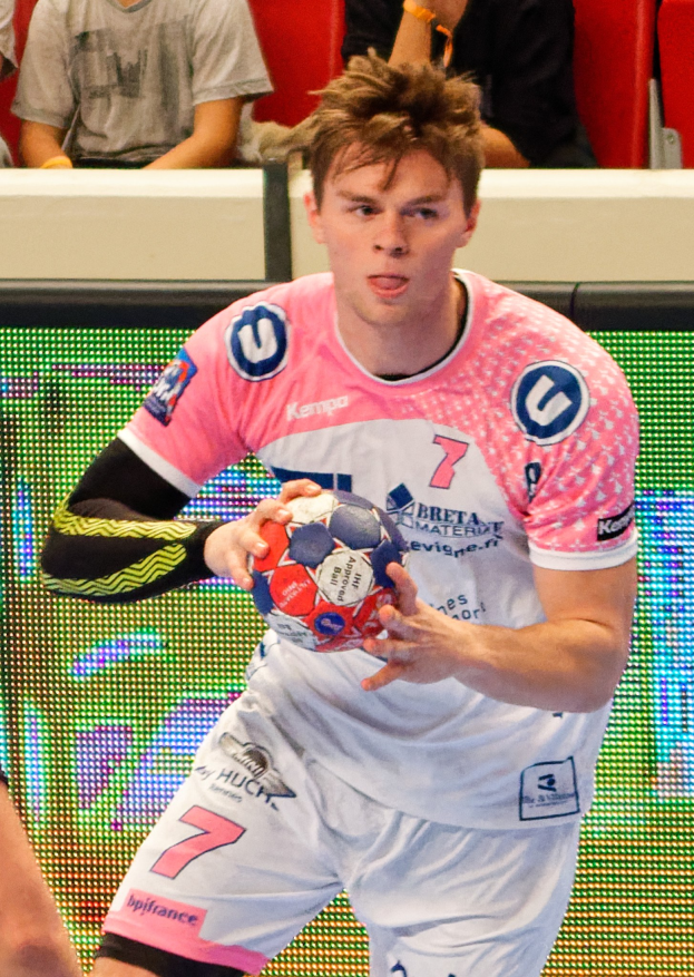 Ein Mann in einem rosa Hemd mit einem Fußball, mit der Hand eines anderen Menschen auf der linken Seite des Bildes und ein paar Menschen im Hintergrund auf Stühlen sitzend.