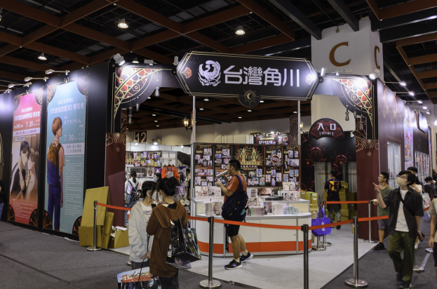 Menschen vor einem Messestand mit Werbematerialien, Taschen und Deckenbeleuchtung, wahrscheinlich auf der Tokyo Anime Expo 2019.