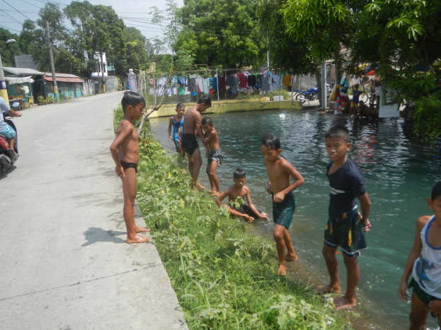 Kinder spielen in flachem Wasser neben einer Straße, einige stehen, andere sitzen auf Gras, mit einer Person auf einem Motorrad links und Häusern, Bäumen und einem klaren blauen Himmel im Hintergrund.