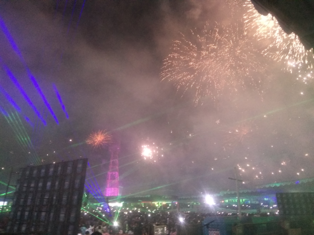 Eine Menschenmenge bei einem Konzert mit beleuchteten Gebäuden und Masten im Hintergrund, unter einem Himmel voller farbiger Feuerwerke.