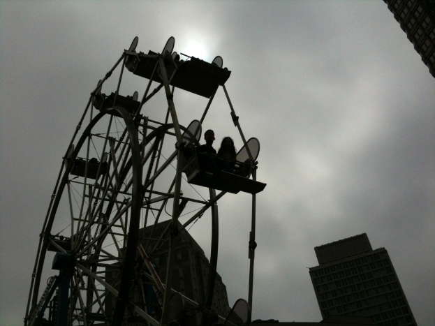 Menschen im Riesenrad mit Gebäuden und bewölktem Himmel im Hintergrund.