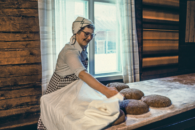 Eine Frau mit Brille und einem strahlenden Lachen hält ein weißes Tuch und steht vor einem Tisch mit Kuchen, mit einer hölzernen Wand, Vorhängen und Fenstern, die Gebäude im Hintergrund zeigen.