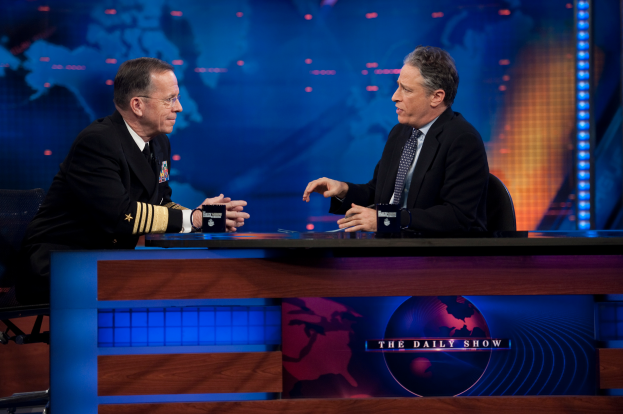 Zwei Manner in schwarzen Blazern, Hemden und Krawatten sitzen an einem Tisch mit Mikrofonen zwischen ihnen, vertieft in ein Gesprach vor einem Fernseher und einem blauen Hintergrund.