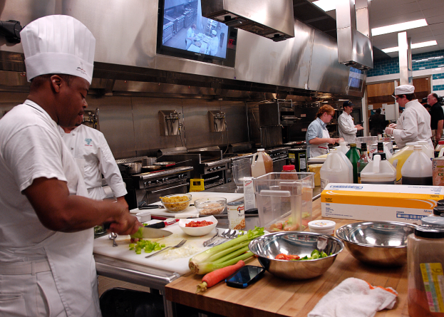Eine Gruppe von Köchen in einer Küche, die Essen auf einem Schneidbrett zubereiten, mit einem Koch in der foreground in einer weißen Schürze und Mütze, der ein Messer hält, umgeben von verschiedenen Utensilien, Schüsseln, Gemüse, einem Mobiltelefon, Flaschen und anderen Gegenständen, mit einem Fernseher und Deckenlampen im Hintergrund.