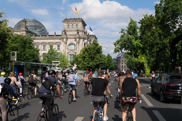 Eine Gruppe von Menschen, die auf Fahrrädern eine von Bäumen gesäumte Straße in Berlin, Deutschland, entlangfahren, mit einer Bushaltestelle auf der rechten Seite und einer Flagge, die über einem Gebäude unter einem bewölktem Himmel weht.