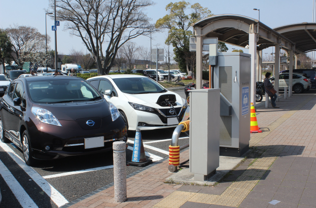 Elektroauto-Ladestation in Japan mit Autos auf der Straße, Verkehrskegel, einer Person auf dem Gehweg, einem Schuppen, Polen, Lampen, Schildern, Bäumen, Pflanzen und einem sichtbaren Himmel.