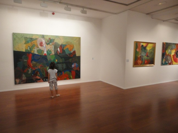 Ein junges Mödchen steht in einer Kunstgalerie und betrachtet gerahmte Bilder an den Wänden, umgeben von anderen Besuchern unter warmem Deckenlicht.