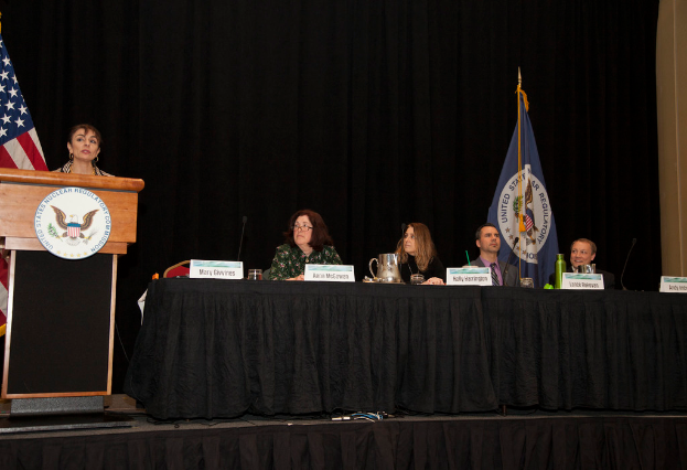 Eine Frau steht an einem Rednerpult auf einer Bühne, spricht in ein Mikrofon, vor ihr ein rundes Schild. Hinter ihr sitzen mehrere Personen an einem Tisch mit Mikrofonen, kleinen Schilden, einer Flasche, einem Glas und einem Krug. Im Hintergrund sind Fahnen, ein Vorhang und eine Wand zu sehen.