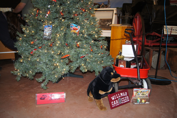 Eine Szene mit einem Hundespielzeug, Namensschildern, einem Einkaufswagen mit Gegenständen, einem Weihnachtsbaum, einem Rad, einem Pfahl, einer Kette, einer Holzplatte, einer Person im Stehen und verschiedenen anderen Gegenständen im Hintergrund.