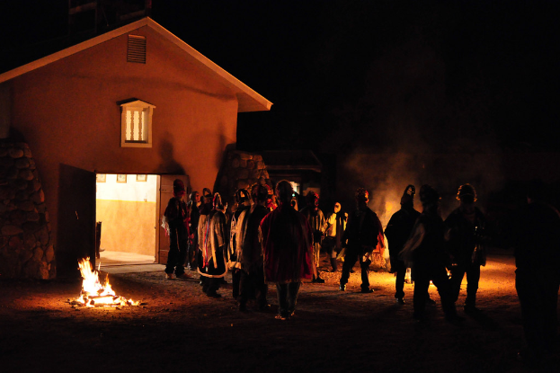 Ein Haus bei Nacht mit ein paar Menschen, die sich um ein Feuer scharen.