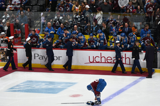 Ein Eishockeyspieler auf dem Eis hält einen Schläger, während er von anderen mit Schlägern und Mützen umgeben ist, mit Zuschauern und Brettern, die "Colorado Avalanche vs San Jose Sharks - 3/24/15 NHL Pick, Odds, and Prediction" anzeigen, im Hintergrund.